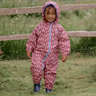 A smiling toddler in an EcoSplash Waterproof Fleece Lined Puddle Suit Pink, standing in a garden with a wooden fence behind.